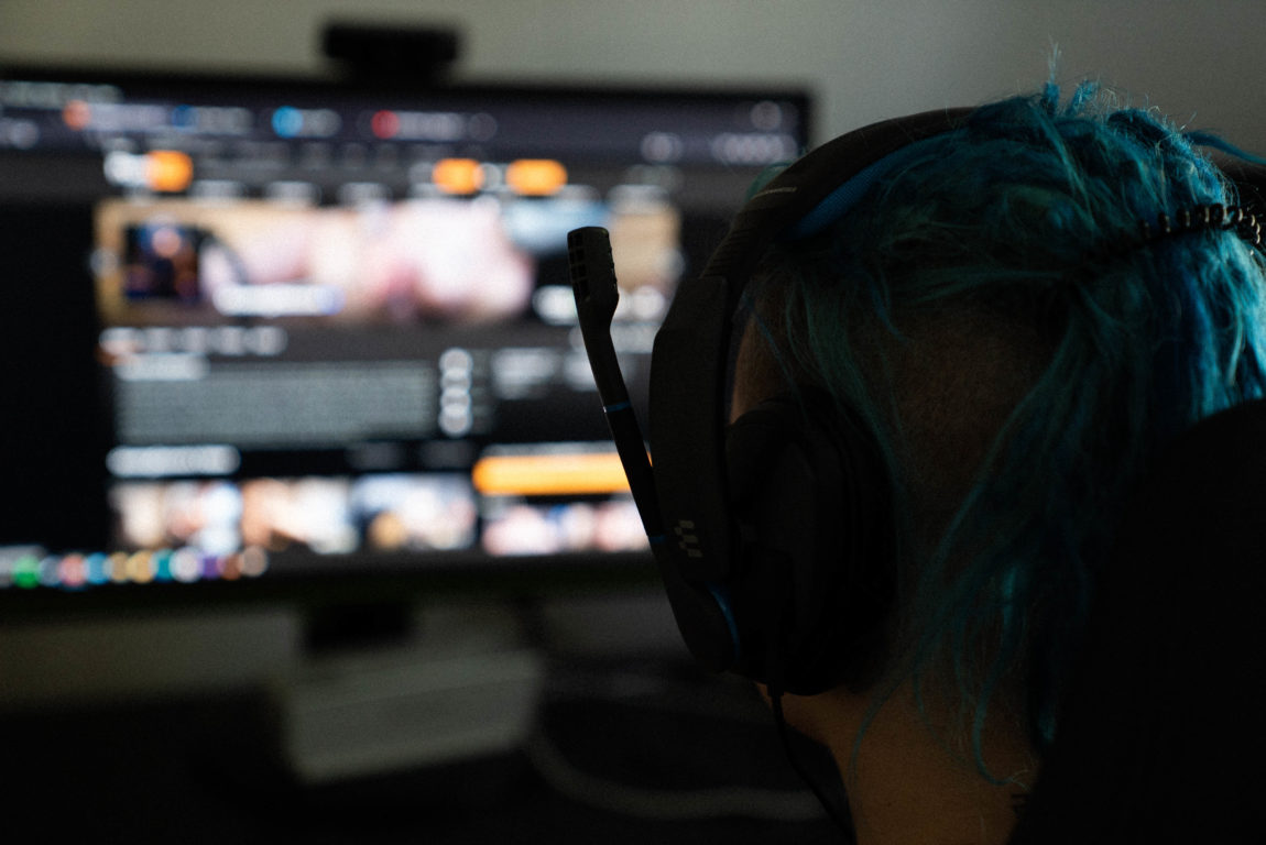 Ei kvinne med blått hår sitt foran ein skjerm med gamingheadset på hovudet. Ein ser berre bakhovudet på kvinna og ein utydeleg skjerm.