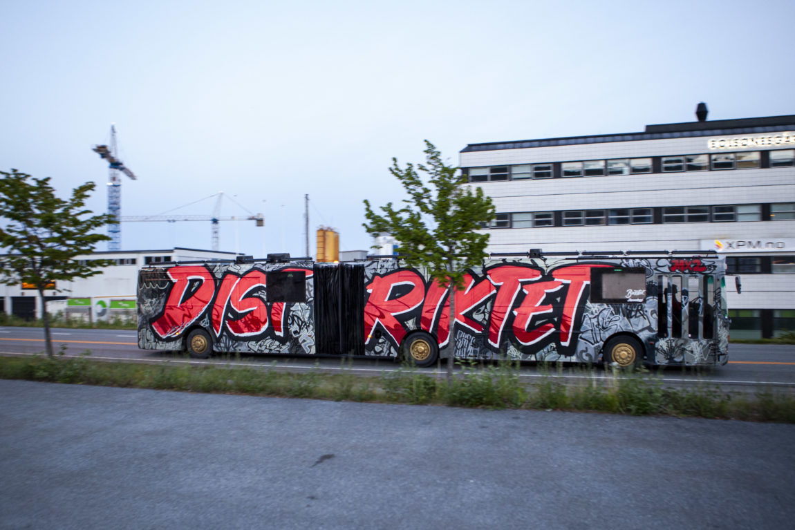 Den lange leddbussen med "Distriktet" malt i godt synleg grafitti, på veg forbi Bolsønes verft i Molde