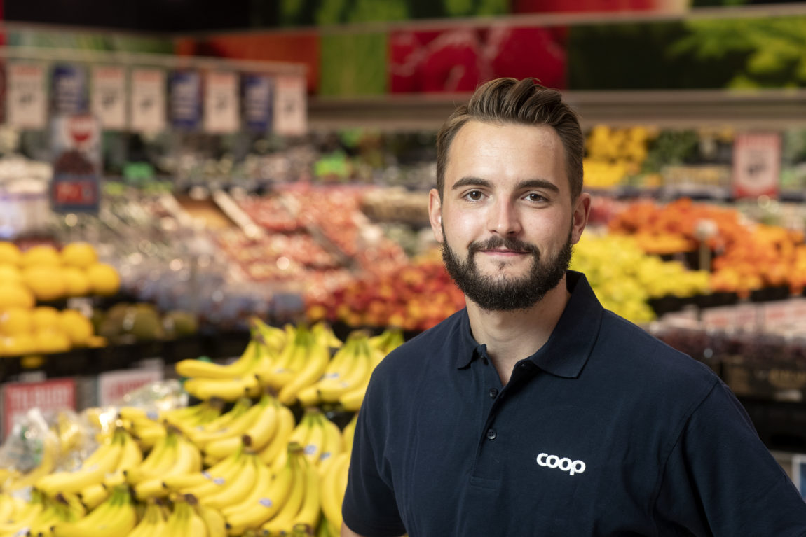 Per Løberg Eriksen har på seg coop t-skjorte. Han står foran frukt og grønt. Han har skjegg, brunt hår og brune øyne. Han har et lurt smil.