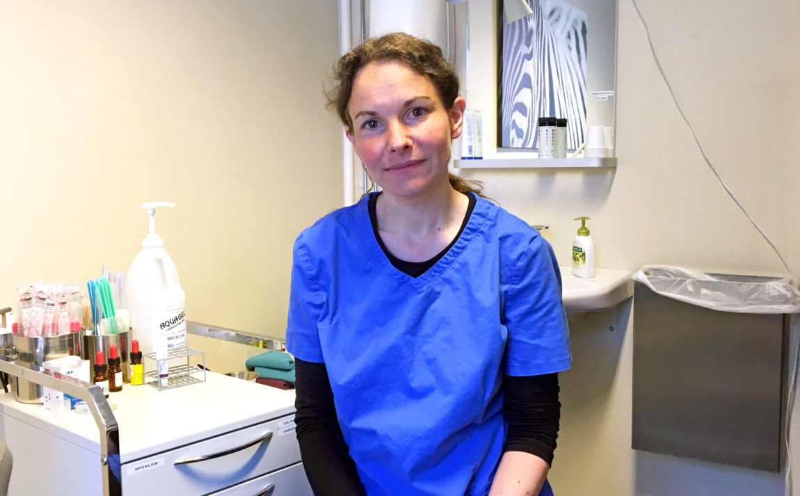 Foto av lege Trine Aarvold. Hun sitter inne på et legekontor og har mørk blå legeskjorte på. Hun ser alvorlig i kameraet, har håret i en hestehale og du kan skimte en vask bak henne og et bord med legeutstyr på til venstre. 