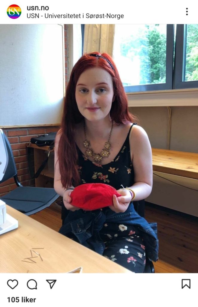 Skjermbilde av Instagrambilde til Universitetet i Sørøst-Norge. Thea ser i kameraet, har rødt langt hår med et hårbånd. På seg har hun er blomstrete kjole og et blomstersmykke hun holder i et rødt munnbind. Hun sitter i et klasserom ved en pult, med vinduer bak seg.