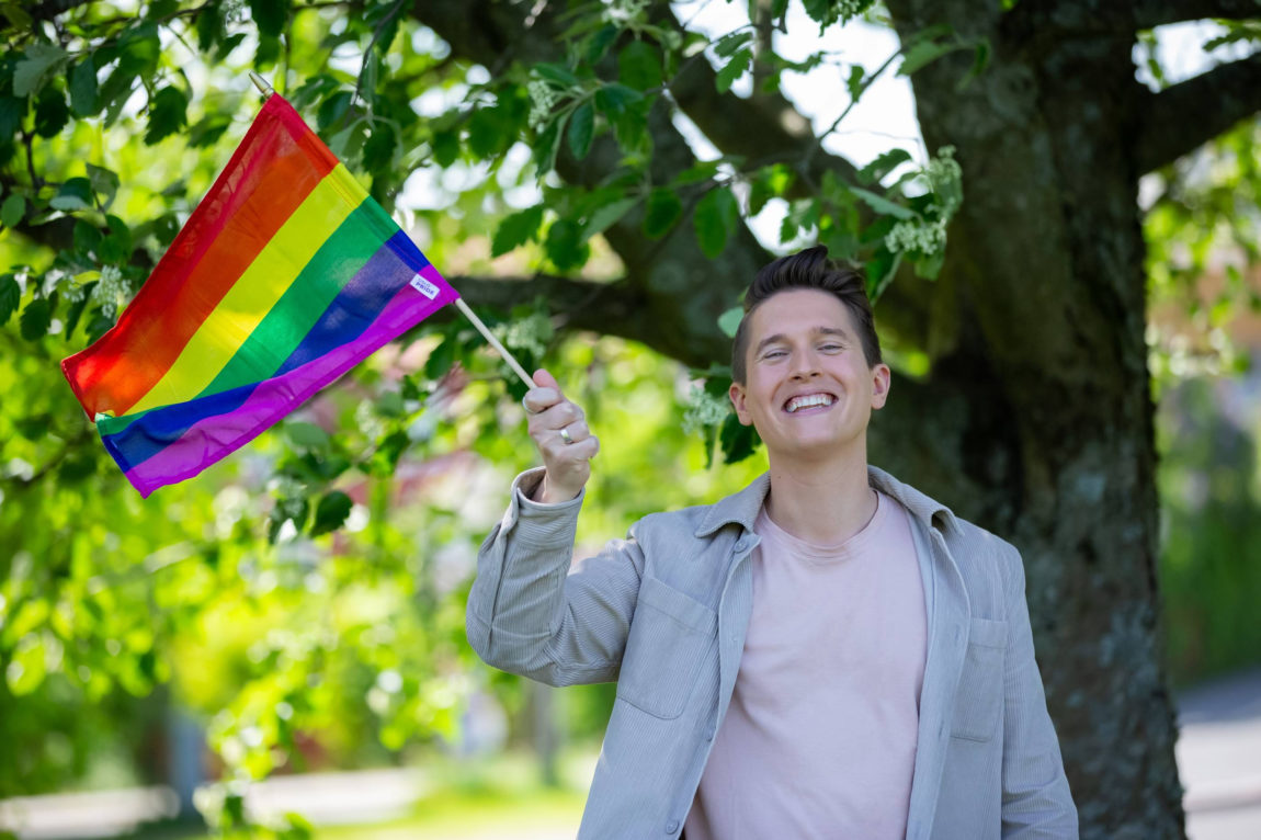 Benjamin har på seg en lys jakke og rosa t-skjorte. Han har et prideflagg i hånden. Han ser inn i kamera og smiler.