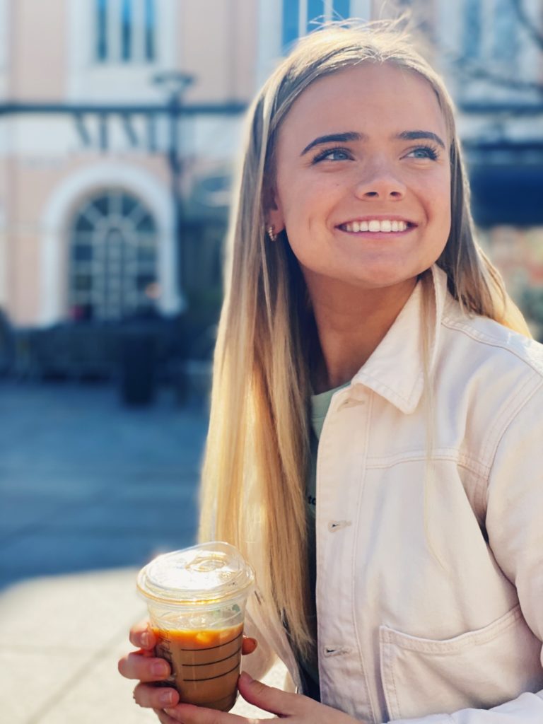 Linnea smiler og ser forbi kameraet. I hånda holder hun en kaffe fra starbucks. Hun har på seg en hvit olajakke og i bakgrunnen får vi se en blury bygning.
