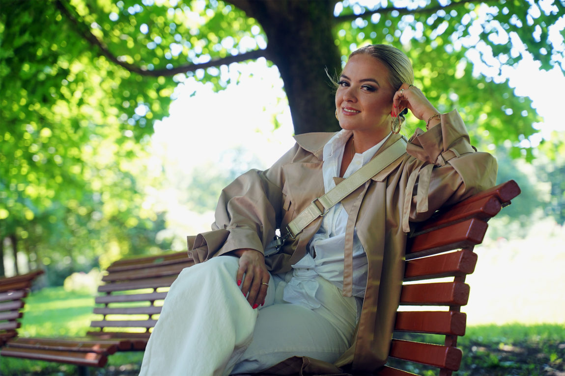 Alexandra sitter på en rød benk i en park. Det er sol og grønne trær bak henne, og hun lener hode på handa. Hun har på seg en hvit skjorte og en beige kåpe med en hvit veske hengende over skulderen. Håret et satt opp i en hestehale.