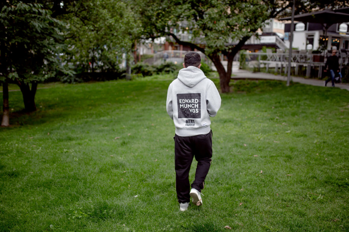 Mert har snudd ryggen til og går gjennom en gresskledd park. På genseren står hans det skrevet «Edvard Munch vgs» i hvite blokkbokstaver.