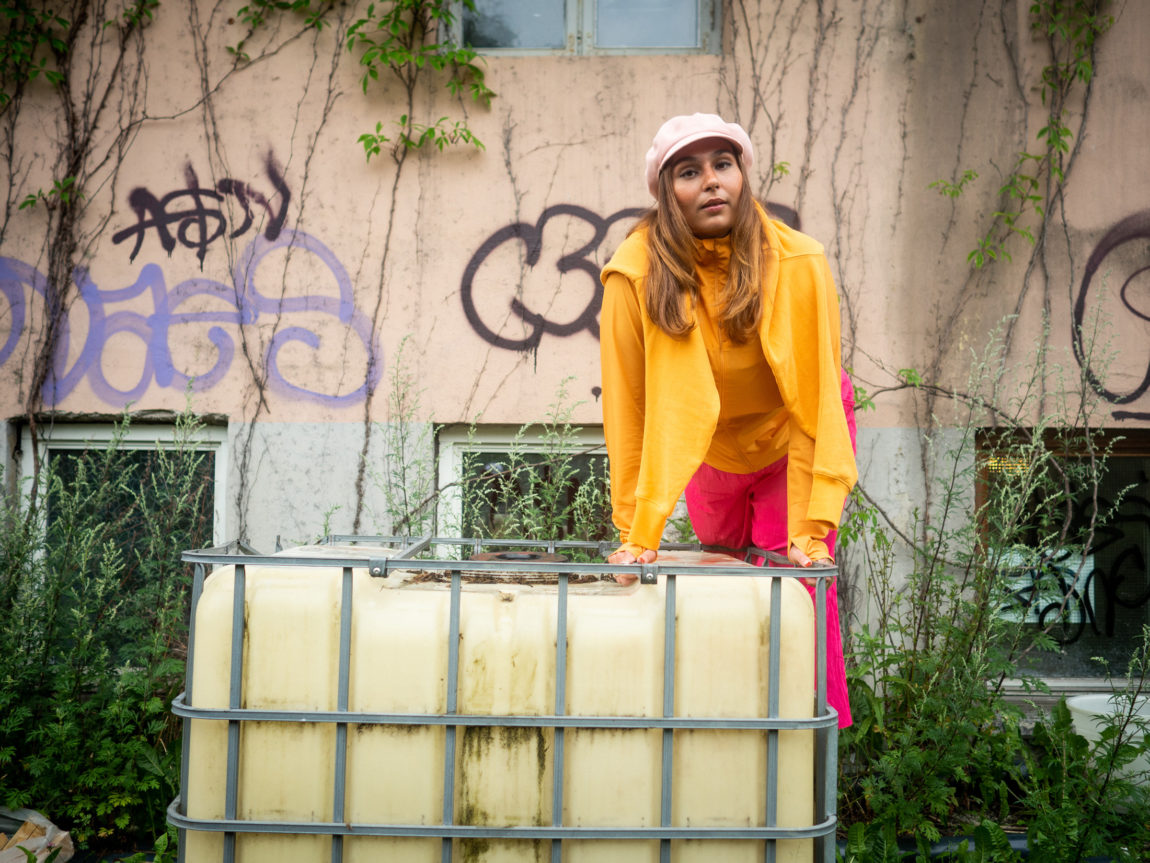 Nedja balanserer lent over en slitt plastboks foran en rosa vegg med klatreplanter og graffiti. Nedja har på seg sterke oransje og rosa klær.