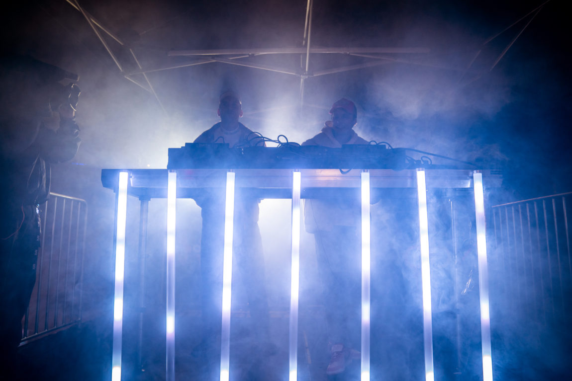 To menn står på en scene, fulldekket i røyk og med blinkende neonlys. 
