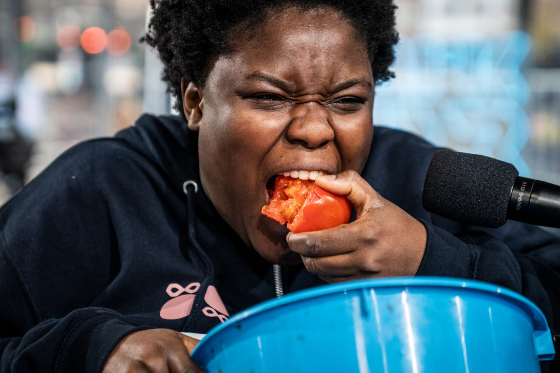 En mørkhudet jente tygger gjennom en svær tomat, mens hun holder en spybøtte foran seg. 