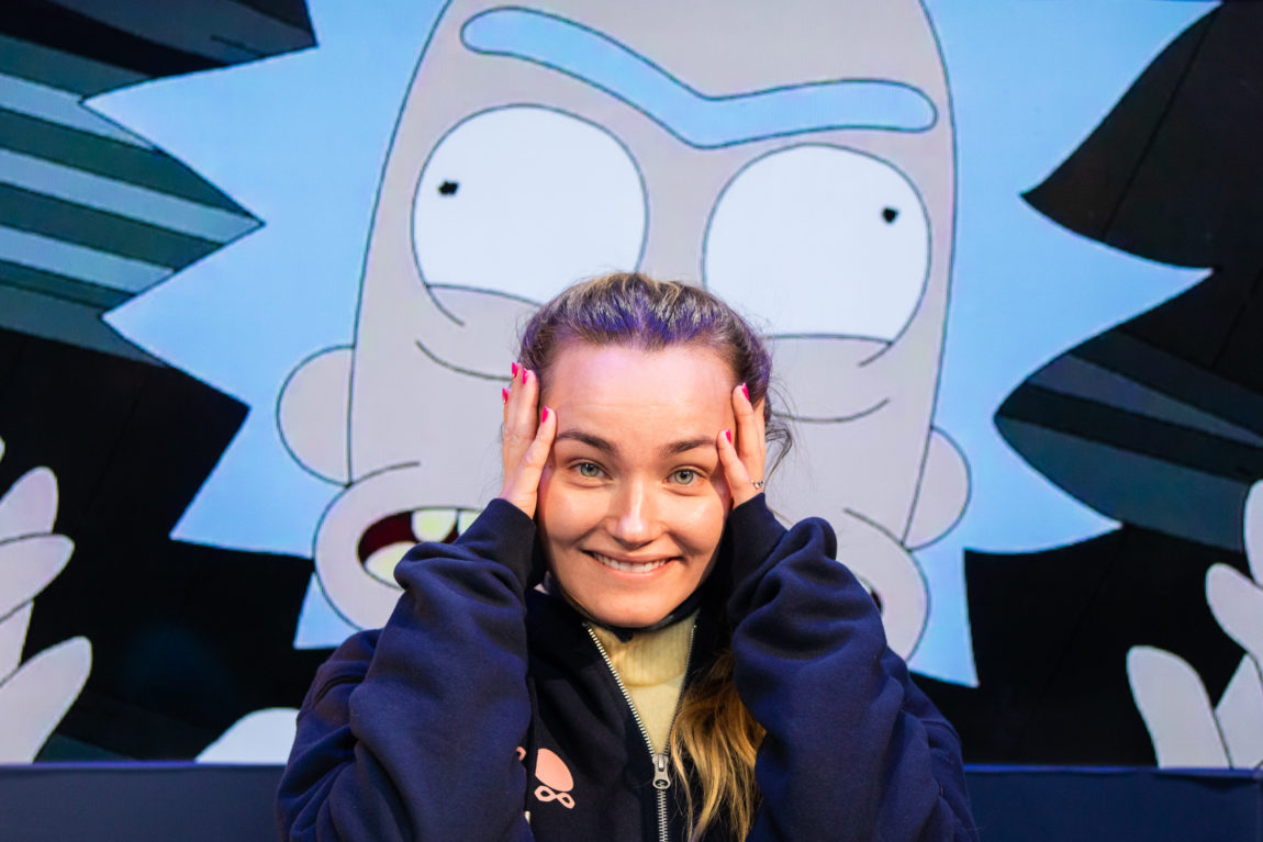 Arian smiler forvirret til kamera, mens hun poserer foran et bilde av en animert, blåhåret karakter. 