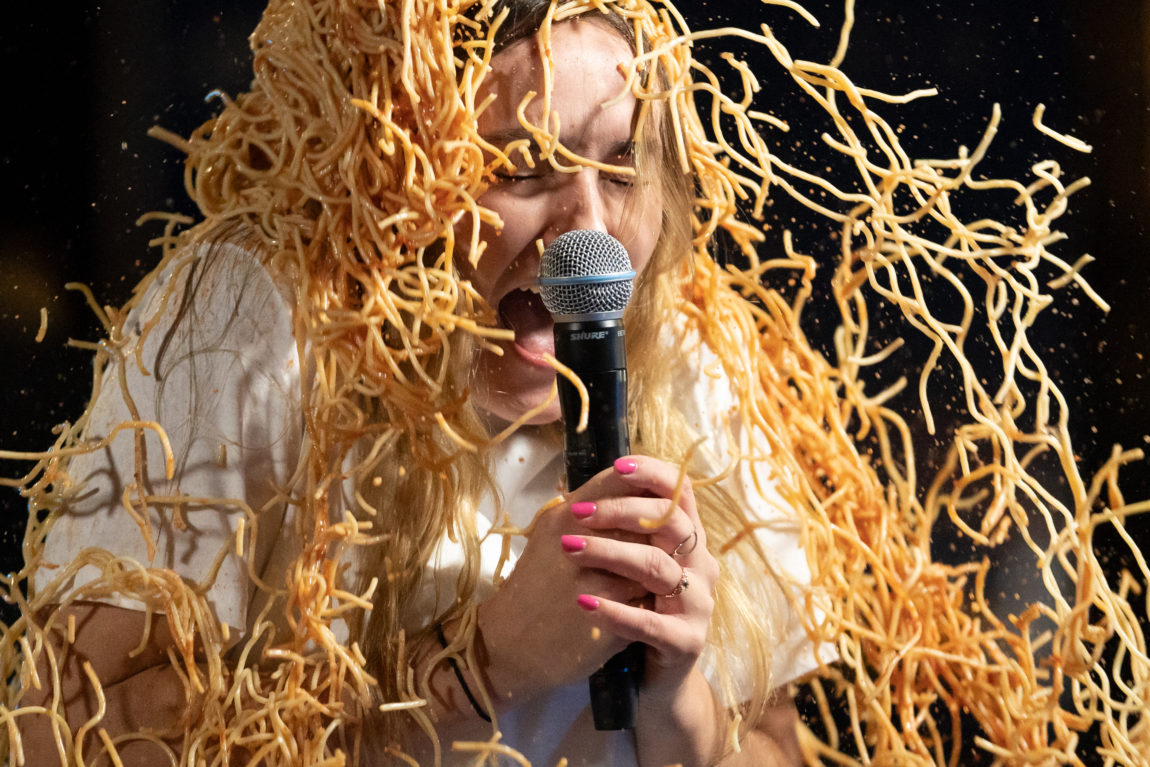Et bilde av en ung kvinne i hvit t-skjorte som holder en mikrofon mens hun skriker av ubehag. En svær porsjon spaghetti med tomatsaus lander i hodet hennes.