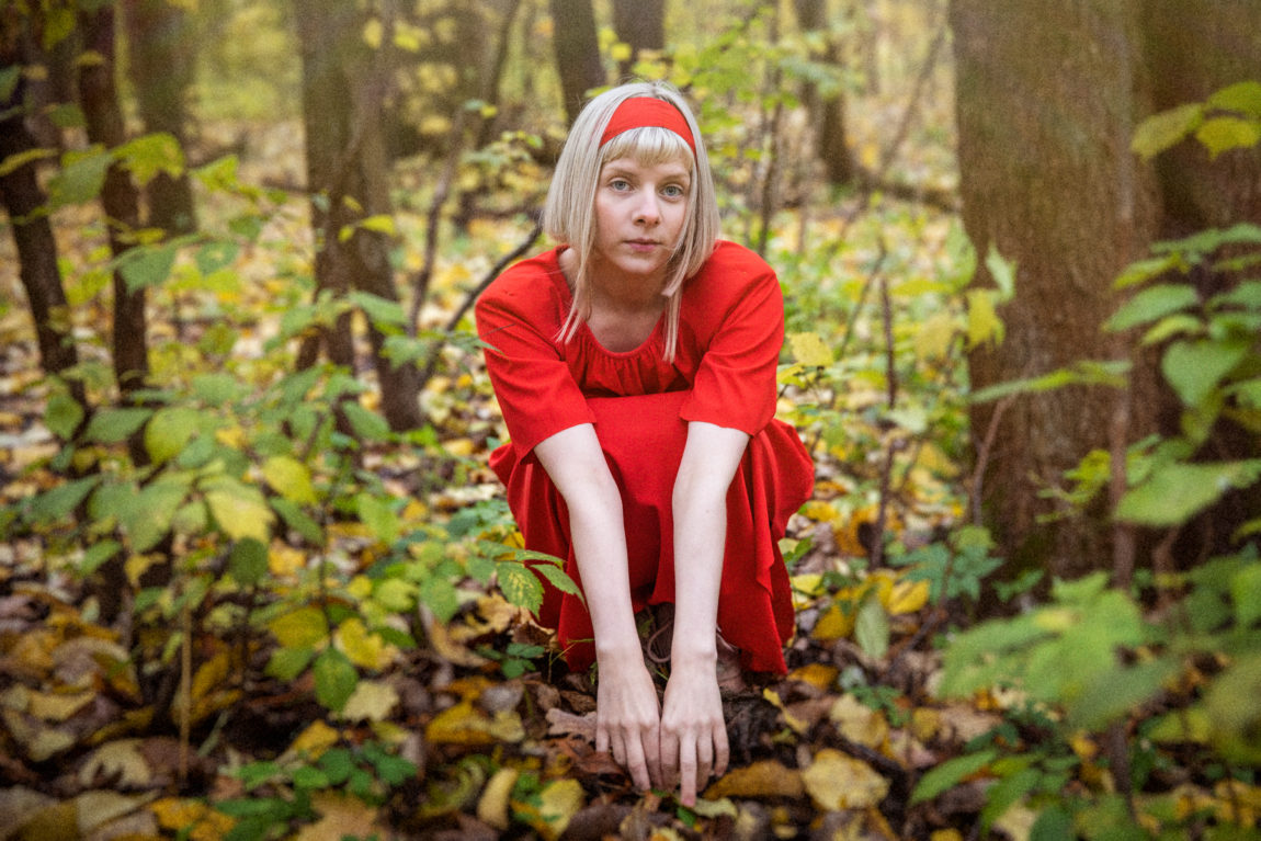 Aurora er ikledd en rød, vid kjole og sitter sammenkrøket med hendene hvilende på knærne, i skogen. Blikket hennes er tilsynelatende alvorlig, men mest avslappet.