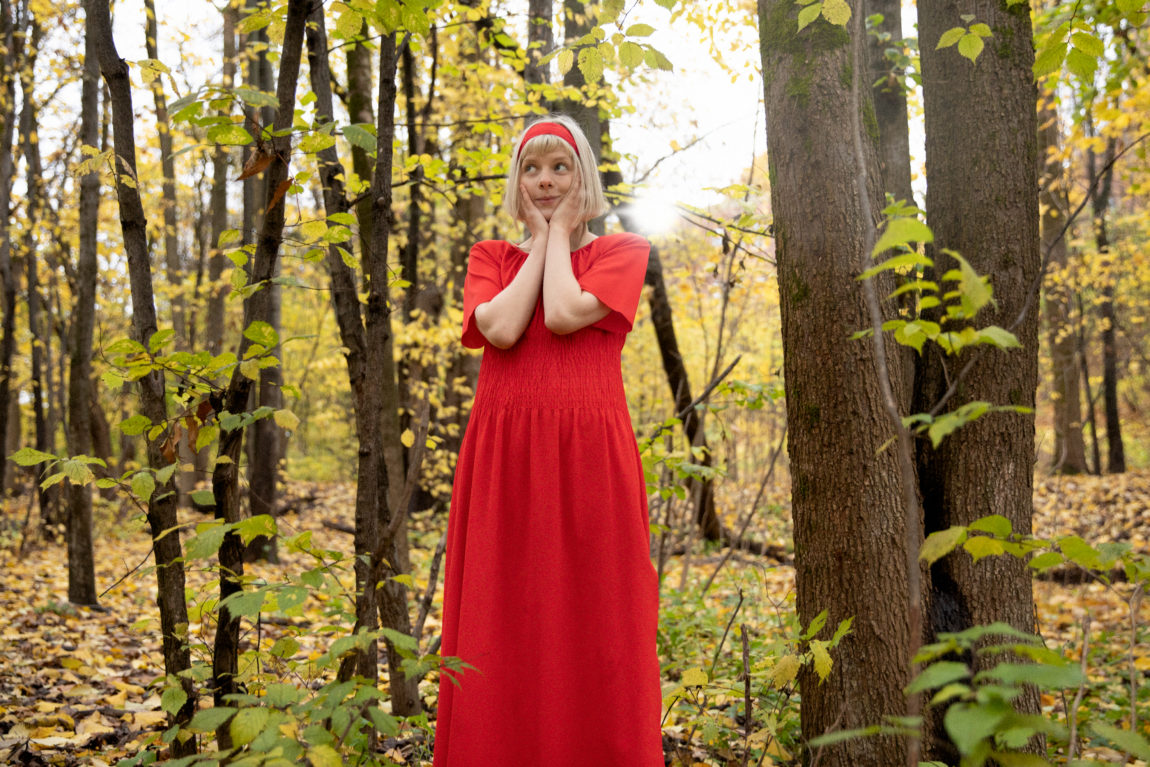 Aurora er ikledd en rød, vid kjole. Hun står i motlys i skogen, og hviler haken tilgjort uskyldig i hendene