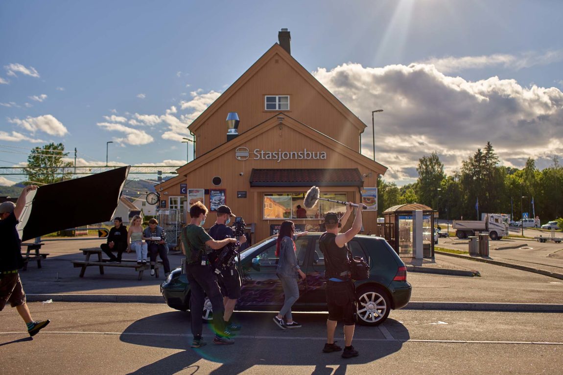 Bilde fra opptak. Kameramann, lydmann og annet crew på bilde. Hege skal inn i en liten mørk bil. Tre stykker sitter på en benk til venstre. Uttafor en togstasjon. Det står stasjonsbua på den oransje byggningen bak bilen. Det er sol og litt skyet. 