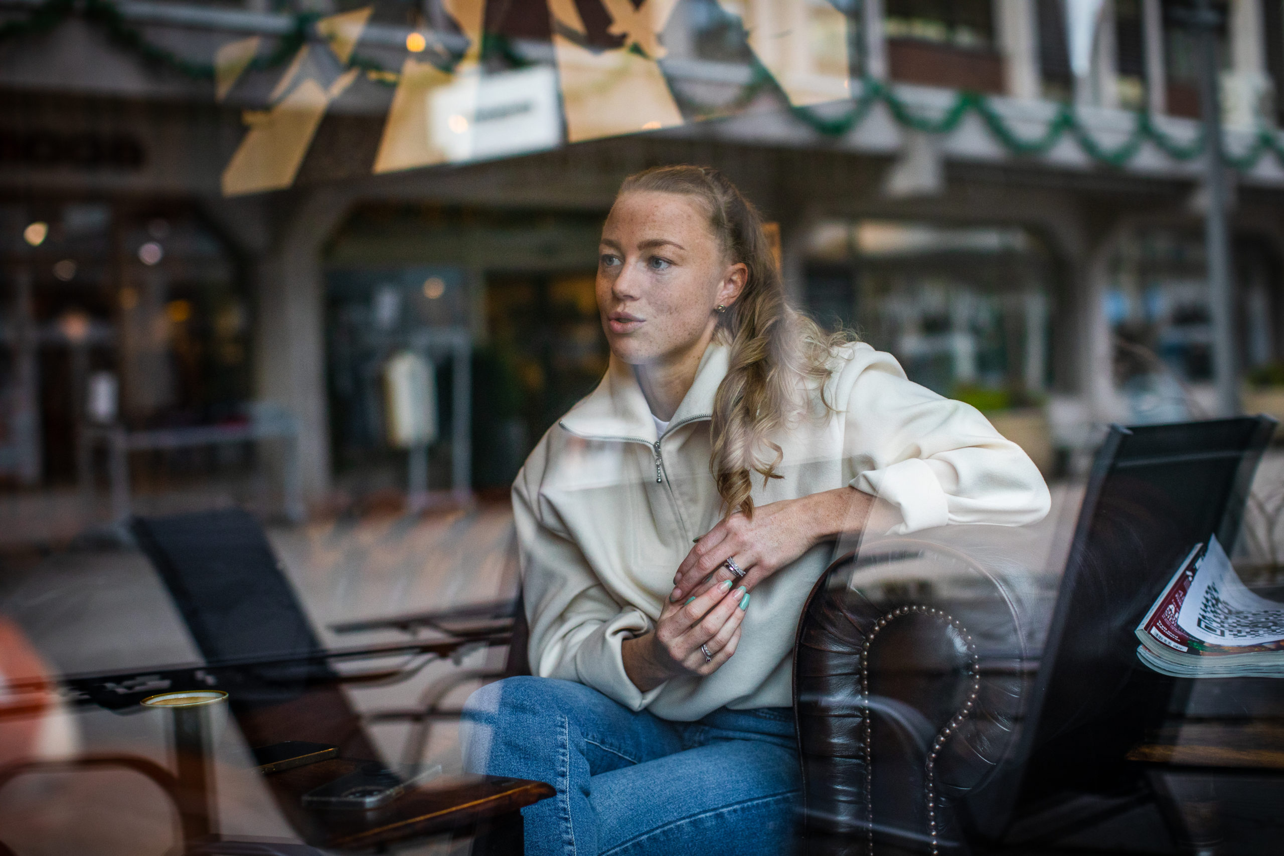 Madeléne sitter framovlent i en skinnstol på en kafe i Fredrikstad. Hun sitter konsentrert og snakker, iført en hvit genser og blå olabuksa.