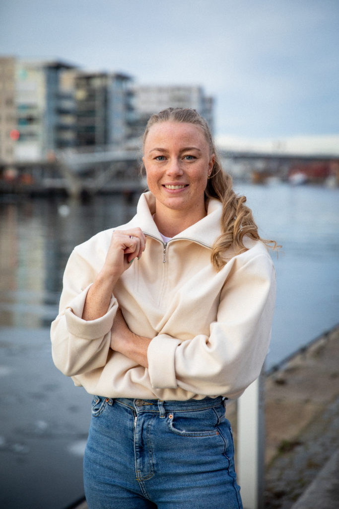 Madelene står foran elvebredden i Fredrikstad og smiler. Hun har håret i hestehale og har på seg en hvit genser og blå olabuksa. 