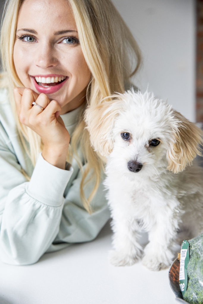 Emilie Nereng sitter ved siden av den lille hvite hunden sin "Eddie". Hun lener hodet i hånden mens hun smiler stort. 