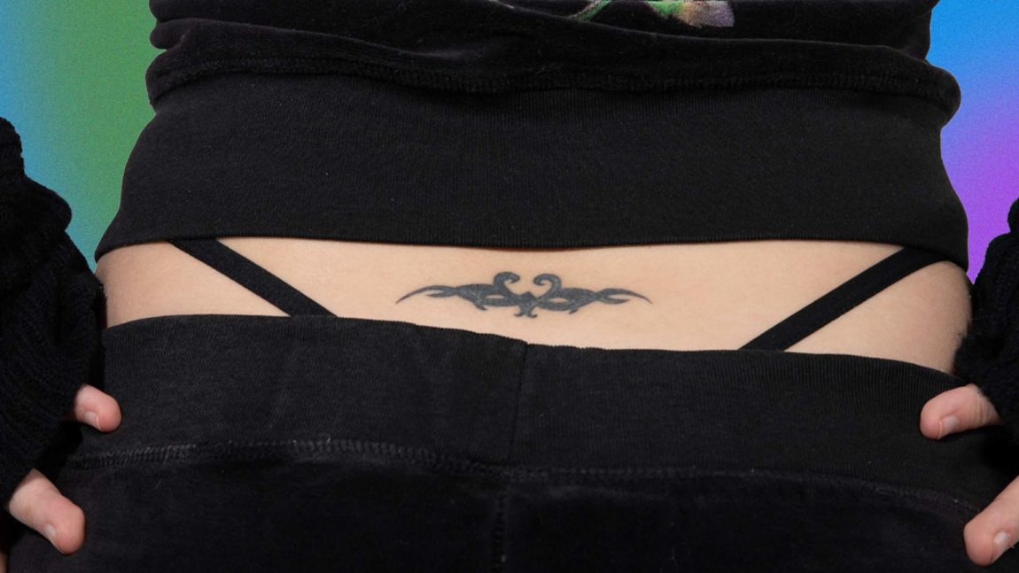 Nærbilde av tatoveringen til Emilie. Det er en såkalt tramp stamp som er på korsryggen. Stringtrusen er synlig på begge sider av hoftene. Hun har på seg et stort antrekk. Hun holder henda på hoftene, to fingre på hver side. 