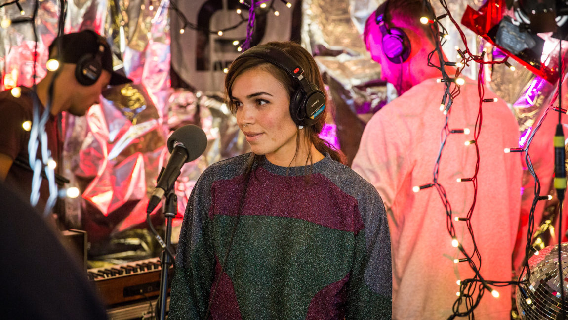 Et bilde av Ary ini et radiostudio. Hun har på seg en fargerik genser i blått, rødt og grønt. Hun smiler mens hun står foran en mikrofon, med hodetelefoner på. Bak henne ser vi to medlemmer av bandet, med hodet vendt vekk fra kamera.