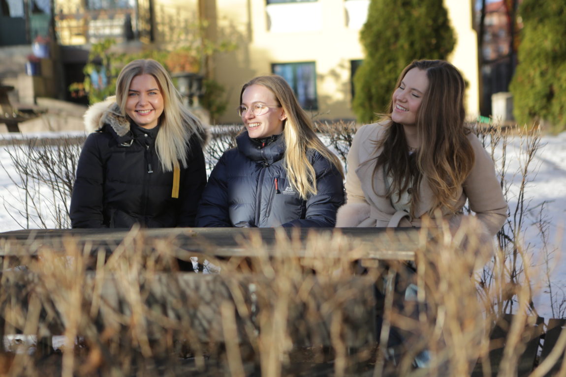 På bildet sitter de tre jentene Selma, Synne og Victoria på en benk. Rundt benken er det lave hekker uten blader på seg, det er snø på bakken og kveldssolen skinner i det gule huset bak dem. 