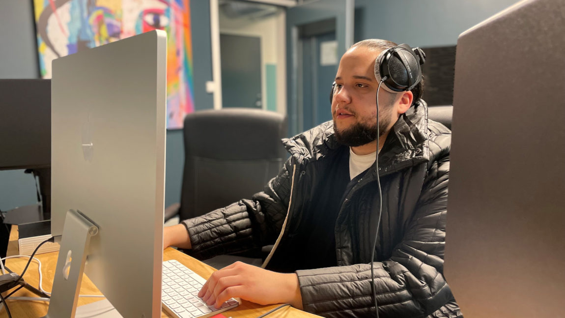 Jonas Benyoub sitter i studio. Vi ser at han jobber. Han har headsettet på hodet. Vi ser bakgrunnen av en pc-skjerm. Han har på seg jakke inne. Har har skjegg.