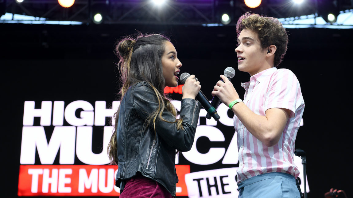 I dette bildet står Joshua og Olivia på scenen med hver sin mikrofon. De ser hverandre inn i øynene mens de synger en duett foran logoen til High School Musical.