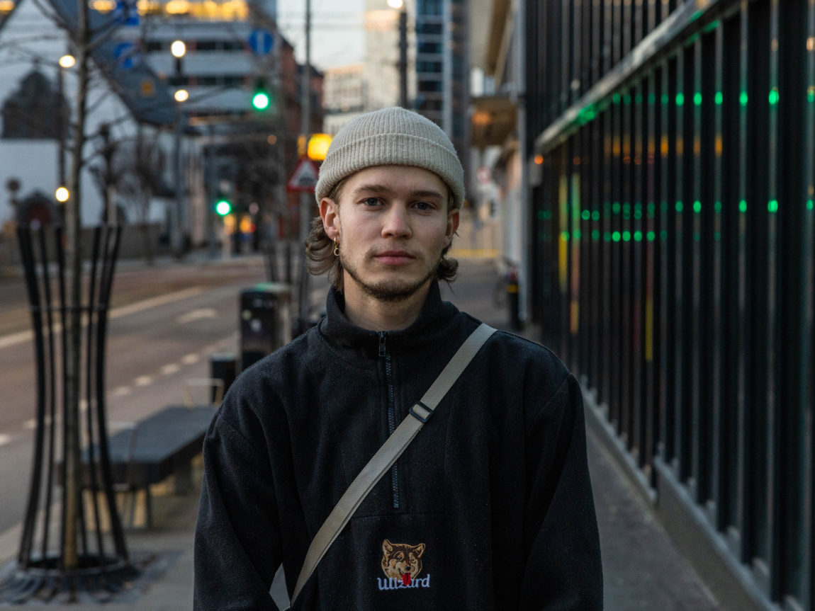 I halvprofil, står André, iført en fleece-jakke og en beanie-lue. Han tilter litt på hodet og stirrer dypt i kameraet. I bakgrunn ser vi gatelys, trafikklys og benker, som indikerer at han er i en stor by. 