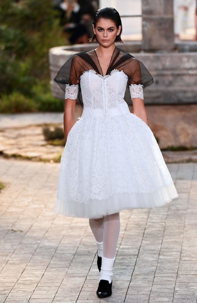 Et bilde av en modell. Hun har på seg en hvit kjole. Det ser ut som hun går på en utendørs catwalk, på brostein.