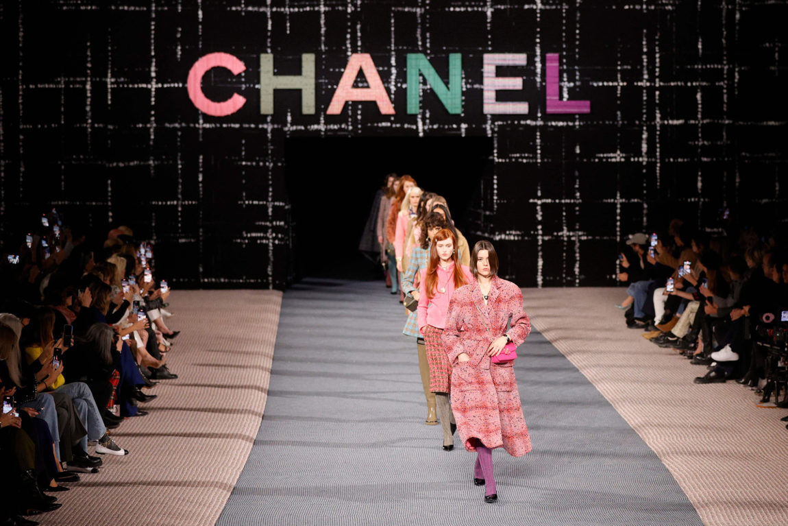 Bildet er tatt i 2022, og viser modeller ikledd høst-vinter kolleksjonen til Chanel for 2022, 2023. Vi kan anta det er slutten av fremvisningen da alle modellene går på rekke over catwalken. Over dem henger fargerike blokkbokstaver som staver "Chanel". 