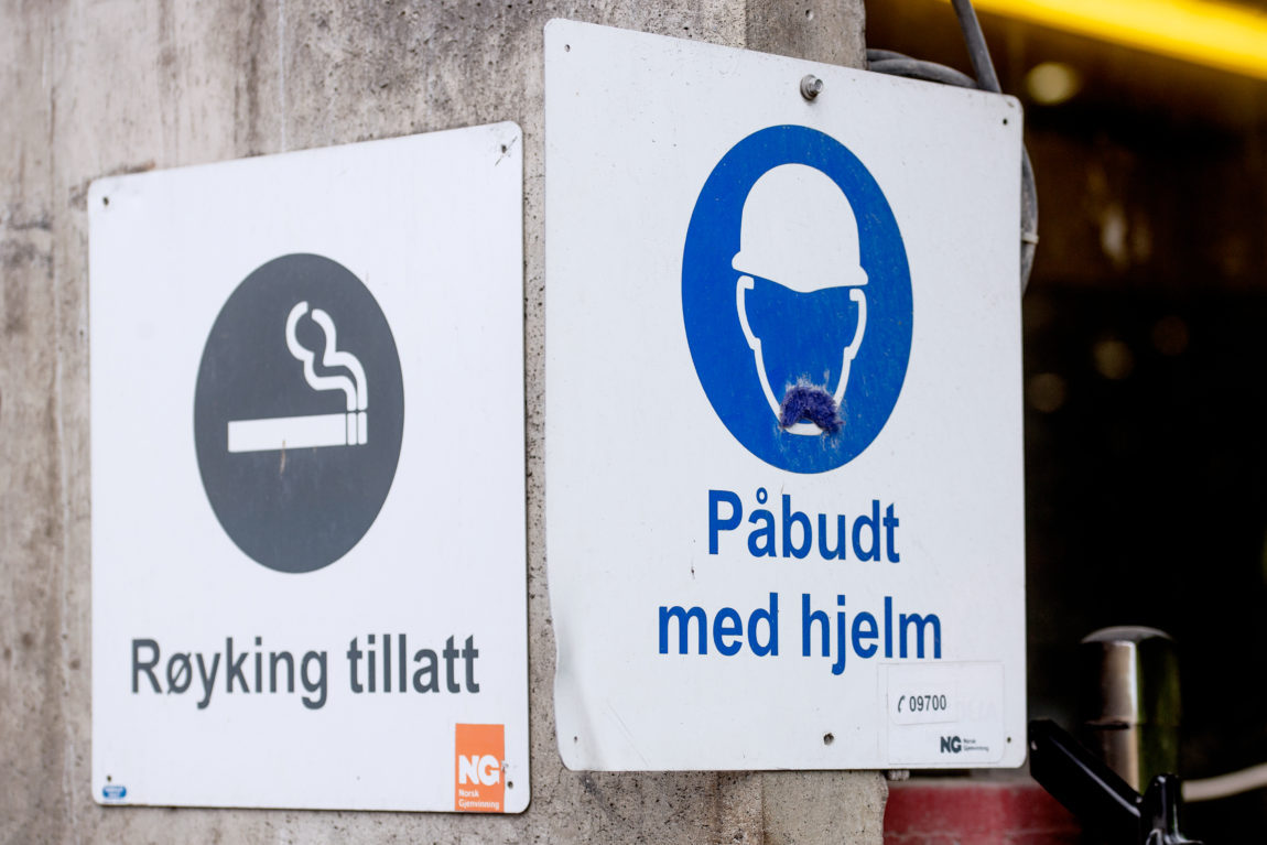 To skilt ved siden av hverandre. På den ene står det «Røyking tillatt»» på den andre står det «Påbudt med hjelm».