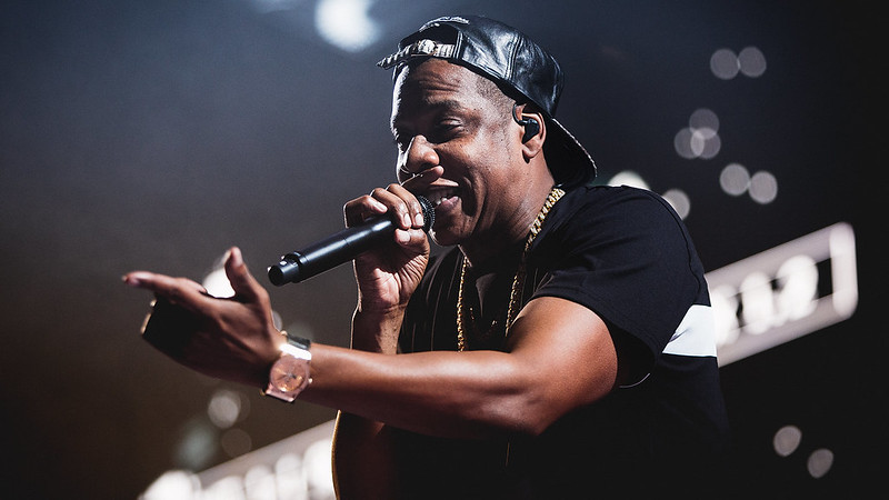 Jay-Z står på scenen med en mikrofon tett mot munnen. Han har på seg sort t-skjorte og sort caps bak frem.