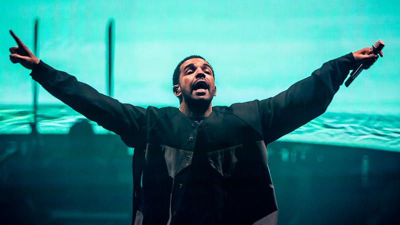 Et bilde av Drake på scenen. Han strekker armene ut til siden mens han holder en mikrofon og roper.