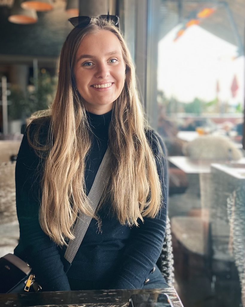 Alexandra Solheim sitter på en restraunt. Hun smiler med tenna og ser inn i kamera. Hun har langt lyst hår og har på seg en sort genser. Hun har en veske på skulderen. Hun har solbriller på hodet. Det er lys bakgrunn.