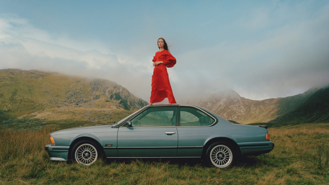 Sigrid har på seg rød kjole og står oppå en blå, gammel bil. I bakgrunnen ser vi fjell og daler under en blå himmel med lett skydekke.