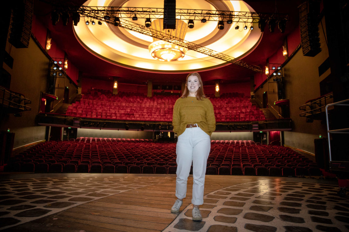 Maren står på scenen. Hun har på seg en sennepsgul genser og gliser bredt. Hun har rødt hår, og bak henne ser vi hundrevis av røde stoler. Der skal publikum sitte snart.