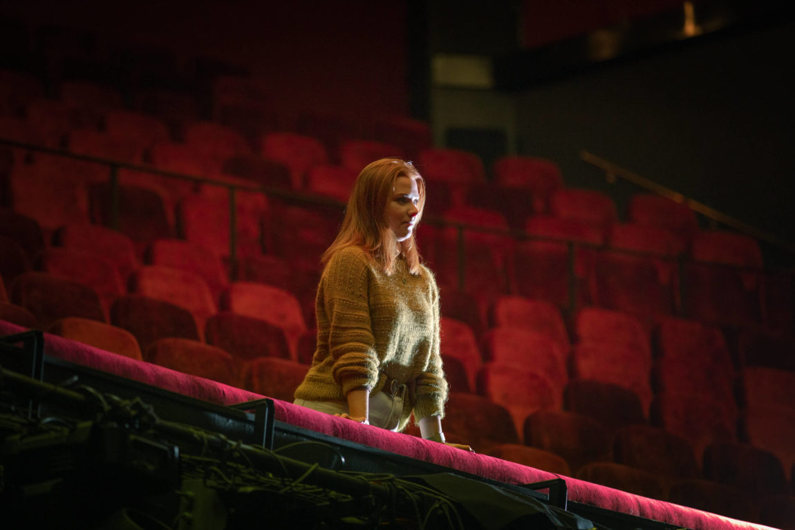 Maren Ovidia står midt på scenen på Folketeateret. Bak henne ser vi 1400 tomme plasser i en dyp rødfarge. Maren har legt hendene bak ryggen, tar et skritt frem og smiler stort. Hun har på seg en grønnbrun strikkagenser og hvit bukse. 