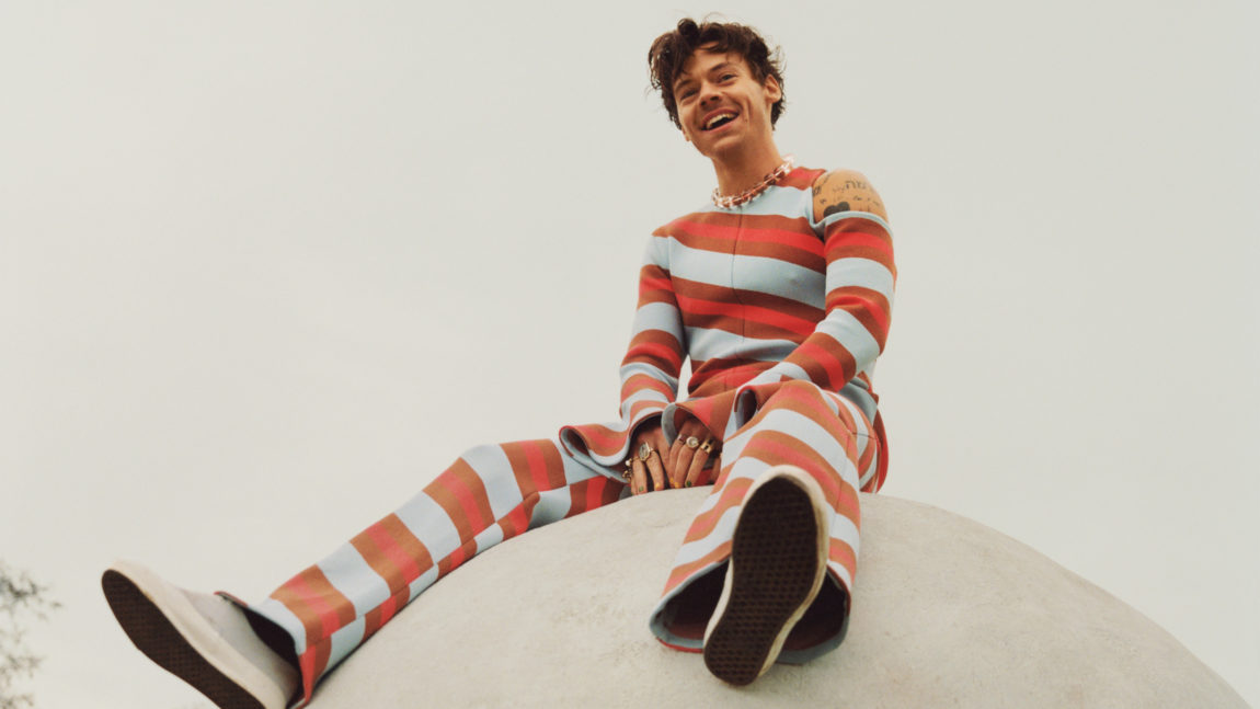 Harry Styles har på seg en onepice med hvite og røde striper. Han sitter på en grå ball.