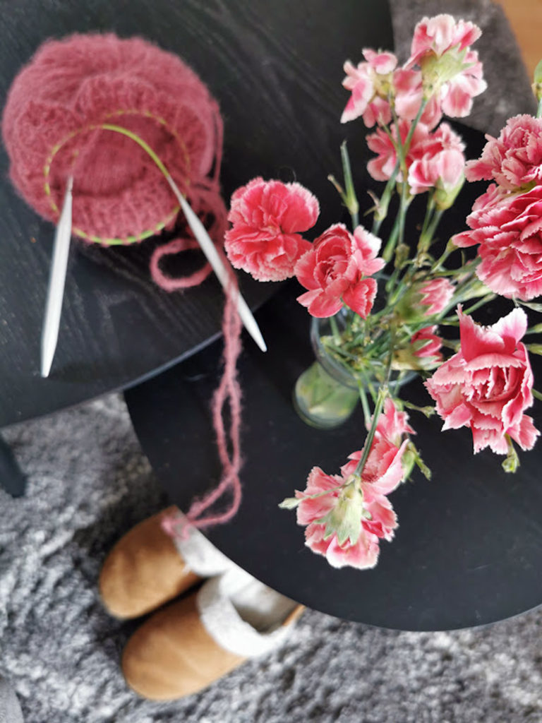Det står rosa blomster i en vase på det sorte bordet som matcher det rosa strikketøyet som ligger ved siden av.