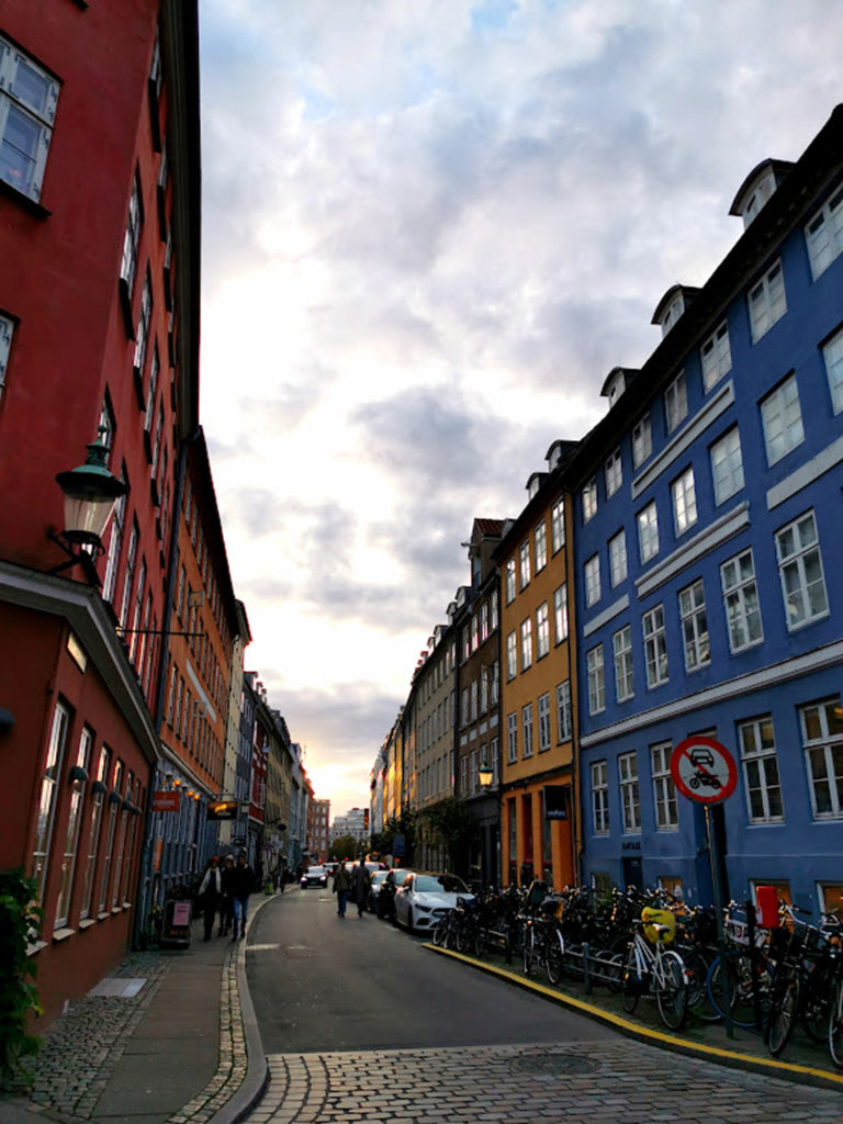 En fargerik sentrumsgate i København. Husene er blå, gule og røde. Himmelen er overskyet og lang veien står det biler og sykler parkert.