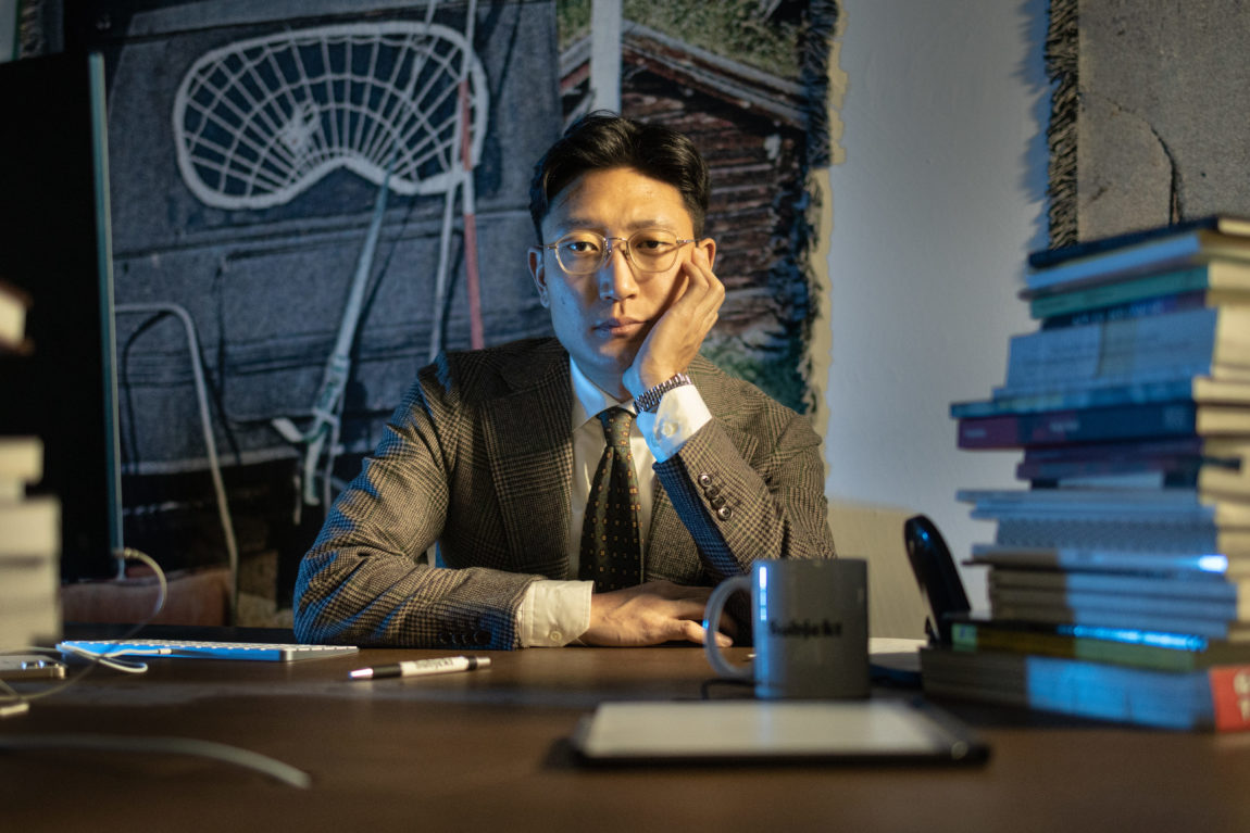 Dandby Choi er en mann med briller, svart hår og rutete blazer. Han sitter på skrivepulten sin og lener hode mot den ene hånden mens han stirrer alvorlig inn i kameraet.