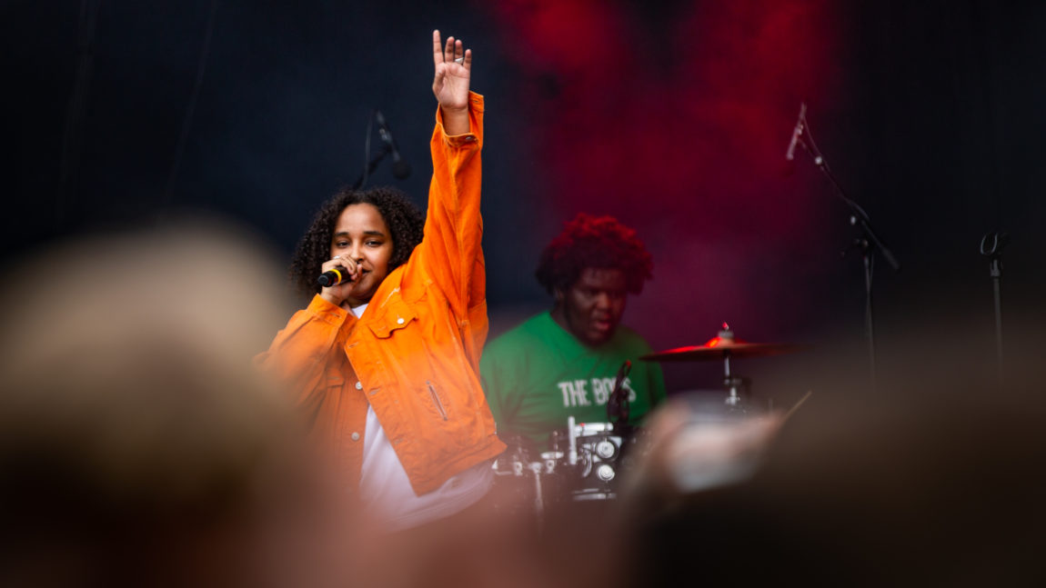 Artisten Musti synger på scenen med en arm opp i været, iført en knall oransje jakke. Bak henne ser vi trommeslageren. 