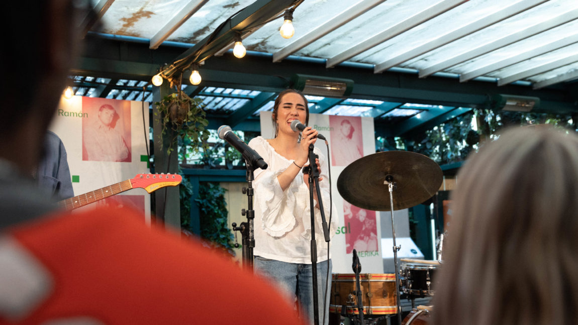 Olivia Lobato har på seg en hvot top og en blå bukse. Hun står på en scene og synger mens hun har øynene igjen. Til høyre er det et trommesett, og til venstre er det en gitar.