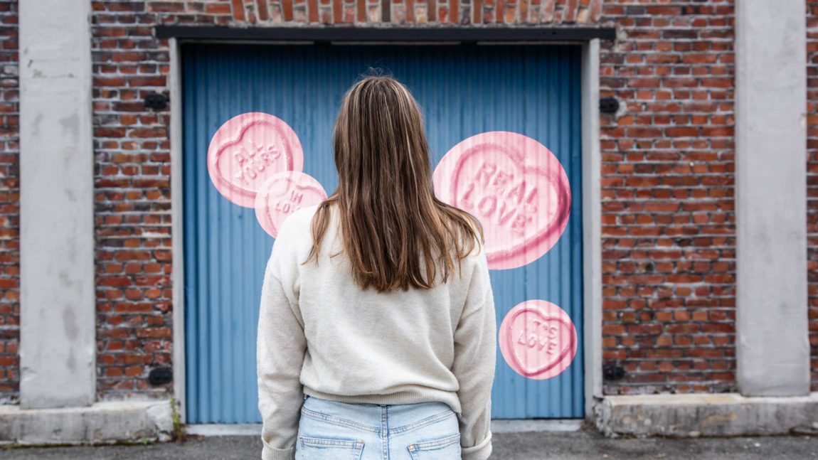 Jamila står framfor ein blå garasjeport, med ryggen til. Ho har på seg blå dongeribukse, kvit gensar og har langt brunt hår. På garasjeporten er det grafisk lagt på "tagging" av rosa lovehearts.