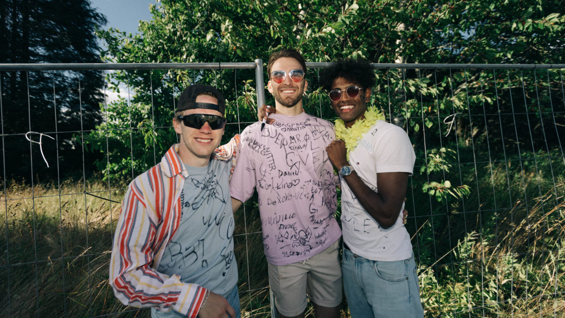 Tre unge menn holder rundt hverandre og smiler. Alle har på seg solbriller og t-skjorter. De har latt andre folk på festivalen har skrevet på t-skjortene, og de ser veldig blide ut. Bak dem er et gjerde og grønne trær. 