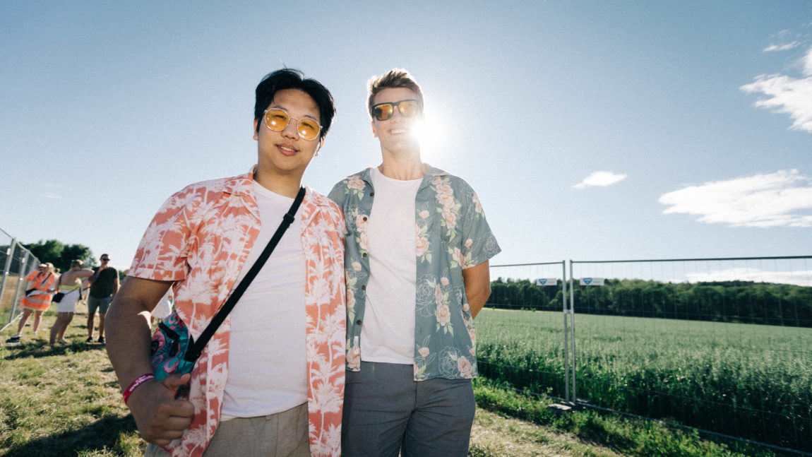 To kompiser som begge har solbriller med gule glass. Han til venstre har en oransje skjorte med hvite busker av et slag, og han til høyre har en blå skjorte med rosa blomster på. 