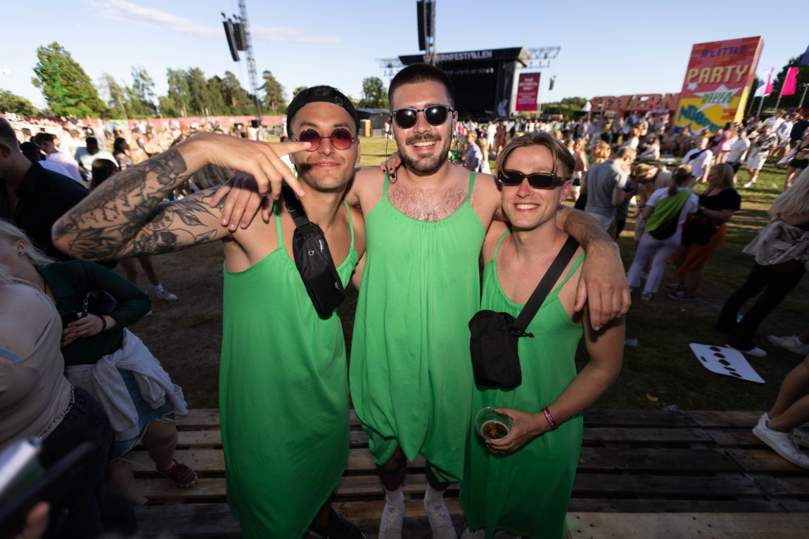 Tre unge menn som har på seg matchende grønne kjoler. Alle har på seg solbriller. Bak dem er det en scene og mange andre festivaldeltakere. 
