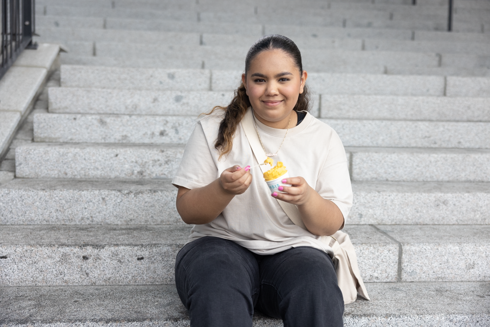 Bilde av Aisha Denne som holder en iskrem i beger i hånden. Hun har brunt langt hår. Hun har på seg en lys t-skjorte og sort bukse. Hun har en lys veske over skulderen. Hun smiler med leppene og ser inn i kamera. Hun har også på et gullkjede.