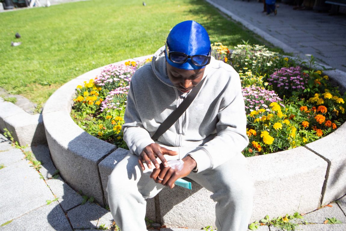 Joel Mambema tar på seg solkrem på hånda. Han har på seg et grå joggedress, durag og solbriller. Han sitter ved et blomsterbed.