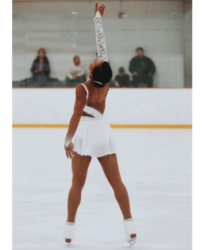 Louisa Warwin på isen. Hun har på seg en hvit kjole og hvite skøyter. Hun står med ryggen til, bena spredd og ene armen opp i været. Uklare mennesker i bakgrunnen.