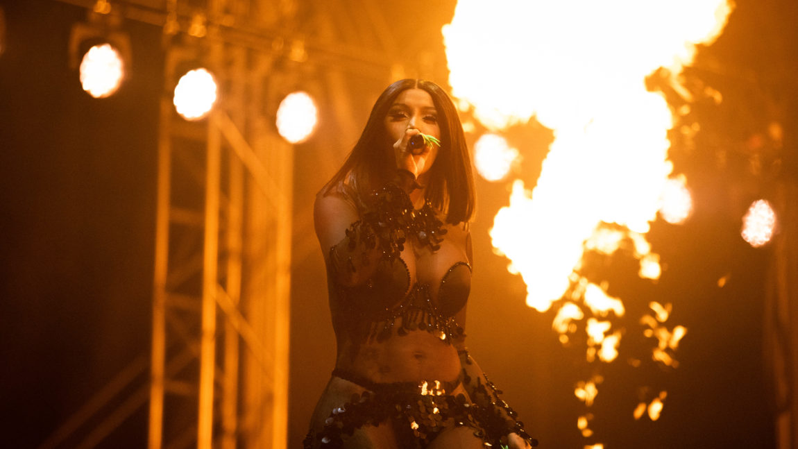 Cardi B har på seg en svart kjole. I bakgrunnen er det flammer. Hun holder mikrofonen tett inntil ansiktet.