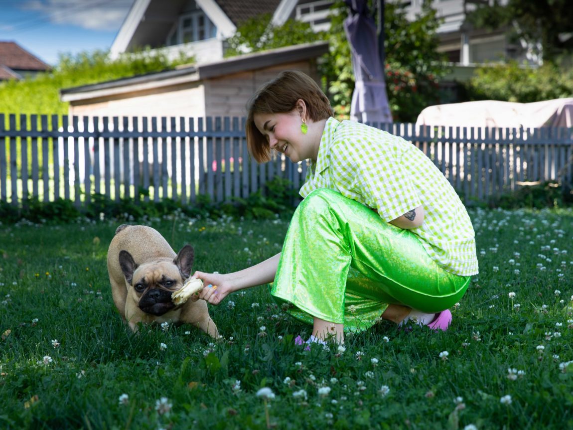 Marie står på huk i hagen og leikar med hunden Fiona. Marie smiler mens ho held ei leike som Fiona bit i. Bak ho er det grønt gras, eit gjerde og nabohus. 